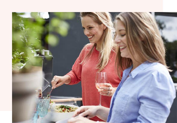 A man and a woman are clinking glasses of red wine together over an outdoor dinner table. They are smiling at each other.