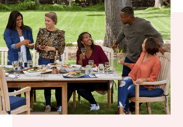 5 people are sitting and standing around a wooden table in a backyard on a sunny day. On the table are plates of food and glasses of white wine.