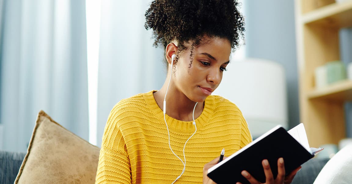 Eine junge Frau sitzt Musik hörend mit einem Notizbuch auf dem Sofa.