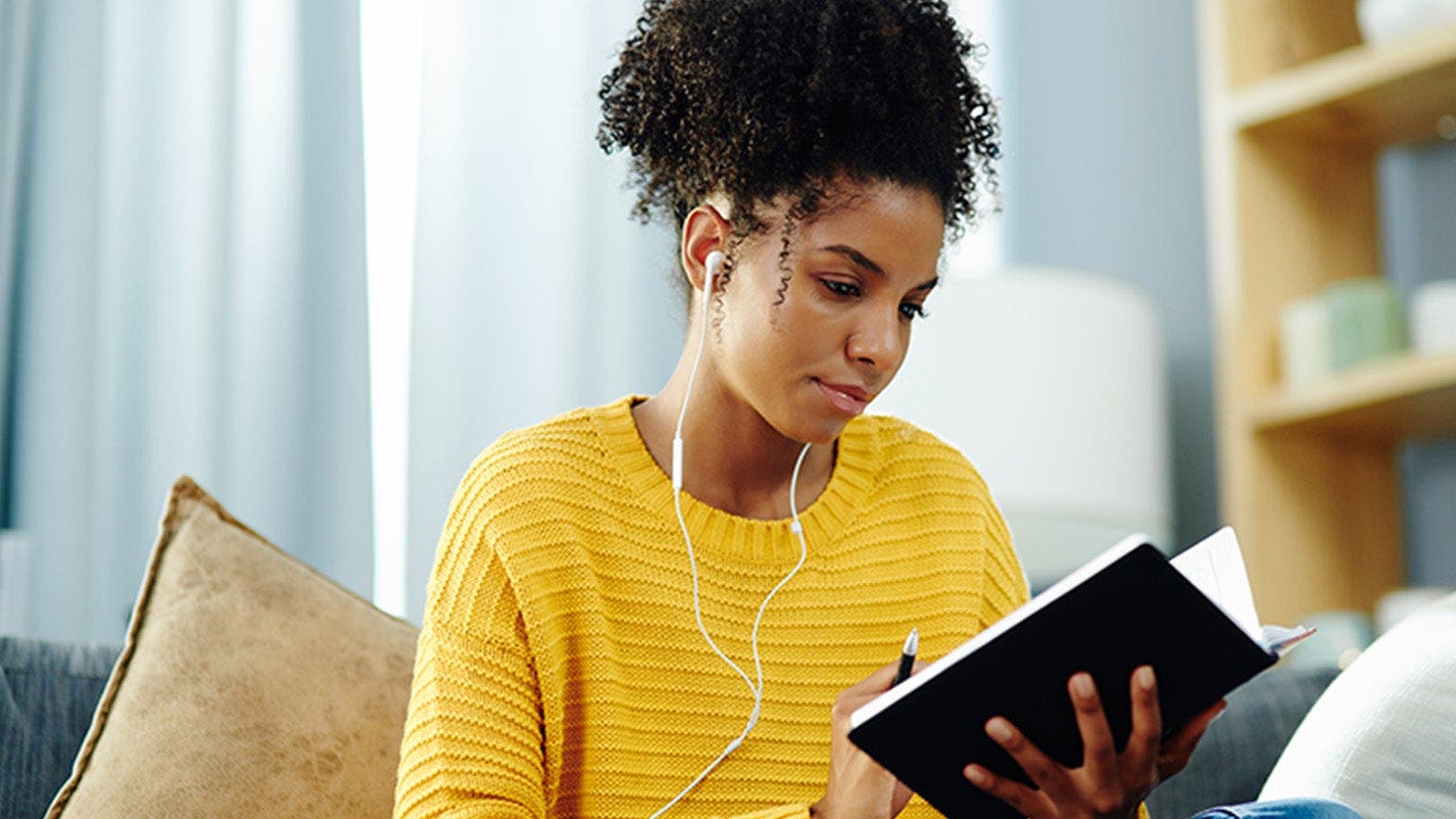 Eine junge Frau sitzt Musik hörend mit einem Notizbuch auf dem Sofa.