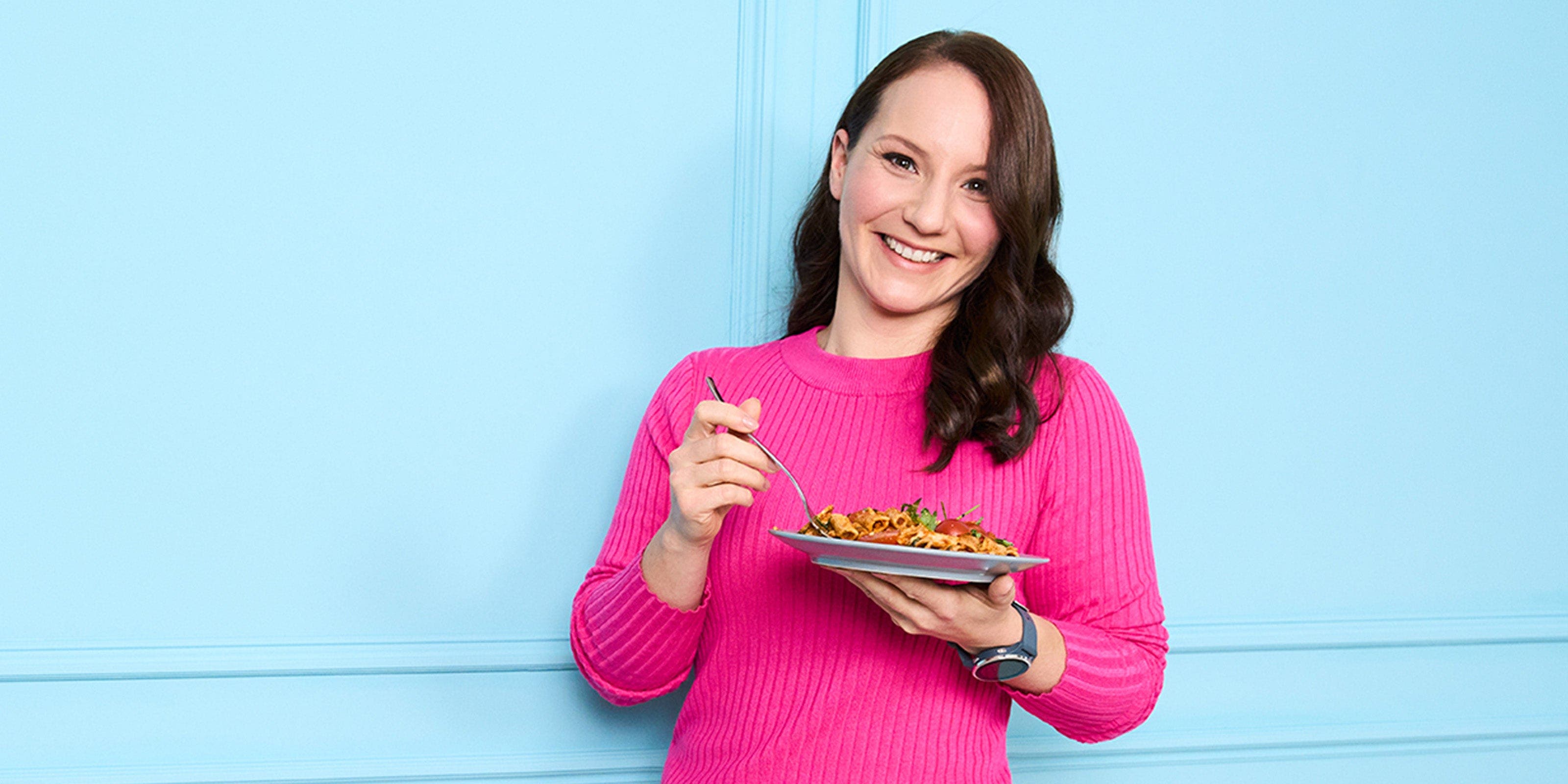 Alexandra, membre de WW, en chemise rose devant un mur bleu, une assiette de pâtes à la main