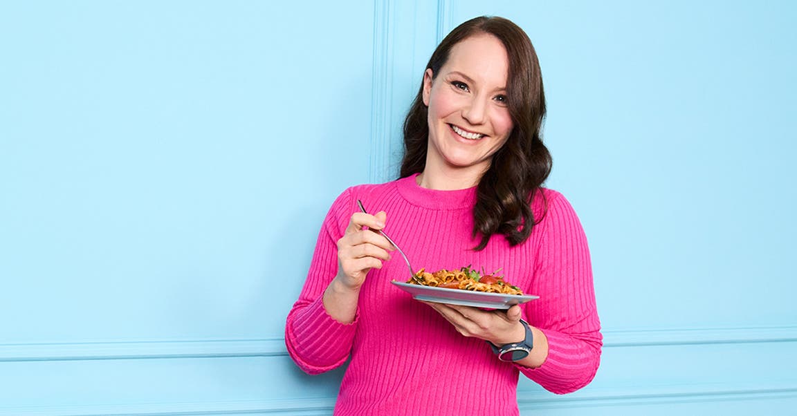 Alexandra, membre de WW, en chemise rose devant un mur bleu, une assiette de pâtes à la main
