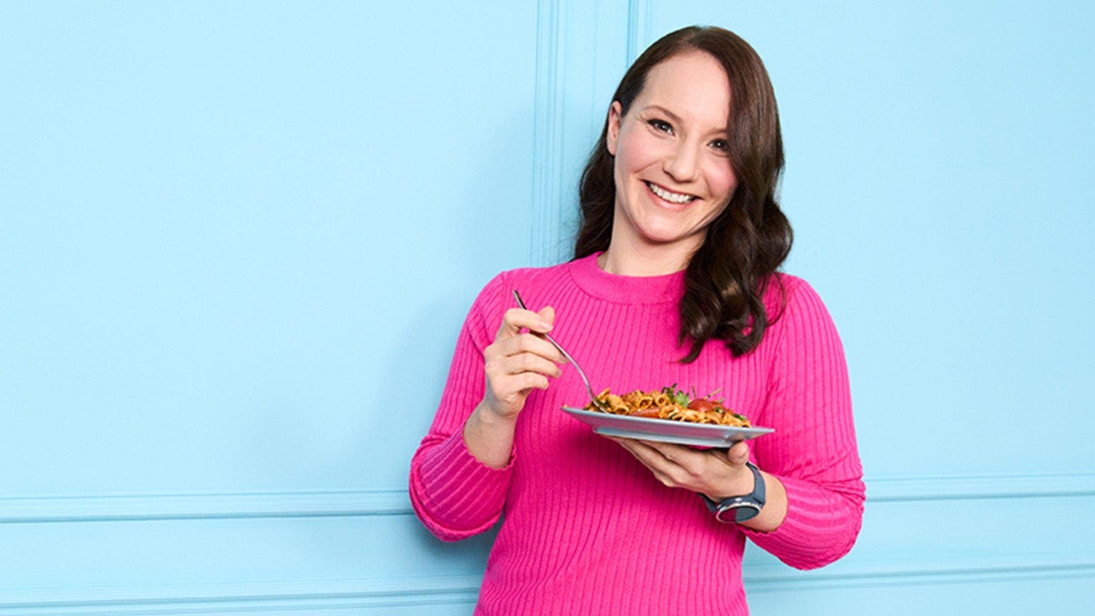 Alexandra, membre de WW, en chemise rose devant un mur bleu, une assiette de pâtes à la main