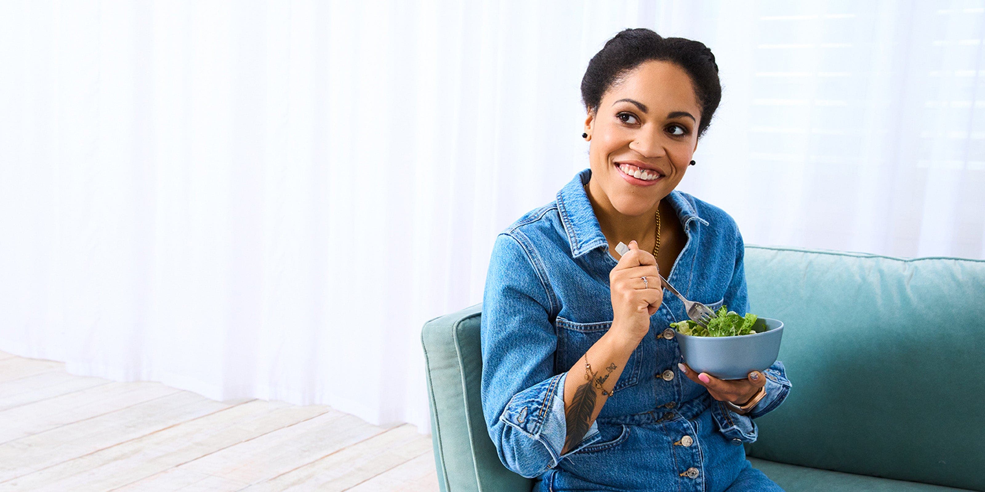 Cecilia, membre de WW, est assise sur un canapé en jean et mange une salade.