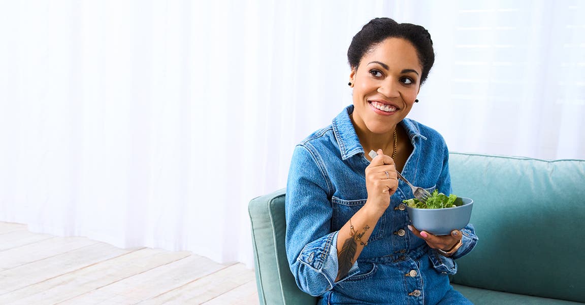 Cecilia, membre de WW, est assise sur un canapé en jean et mange une salade.
