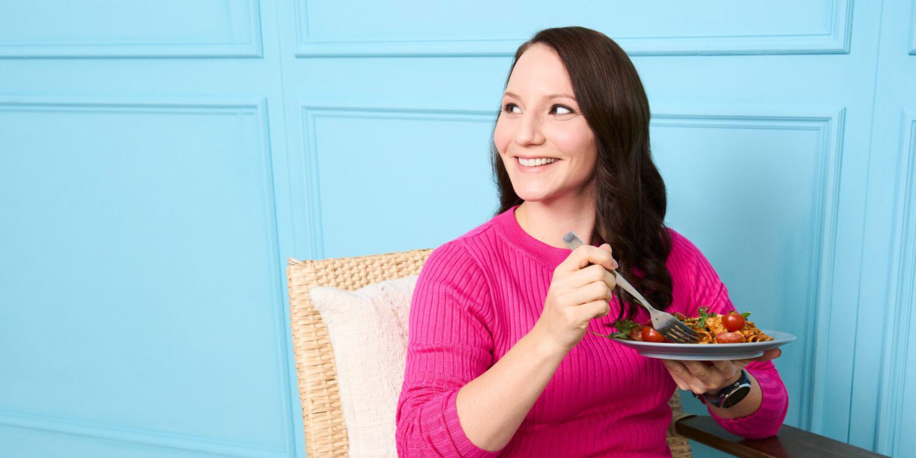 Jeune femme aux cheveux bruns et au pull rose assise avec une assiette de pâtes devant un mur bleu.