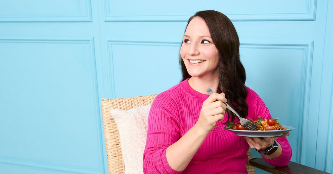 Junge Frau mit braunen Haaren und pinkem Pullover sitzt mit Pastateller vor blauer Wand.