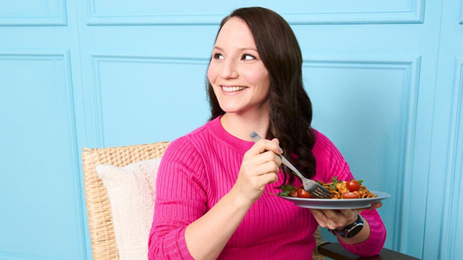 Jeune femme aux cheveux bruns et au pull rose assise avec une assiette de pâtes devant un mur bleu.