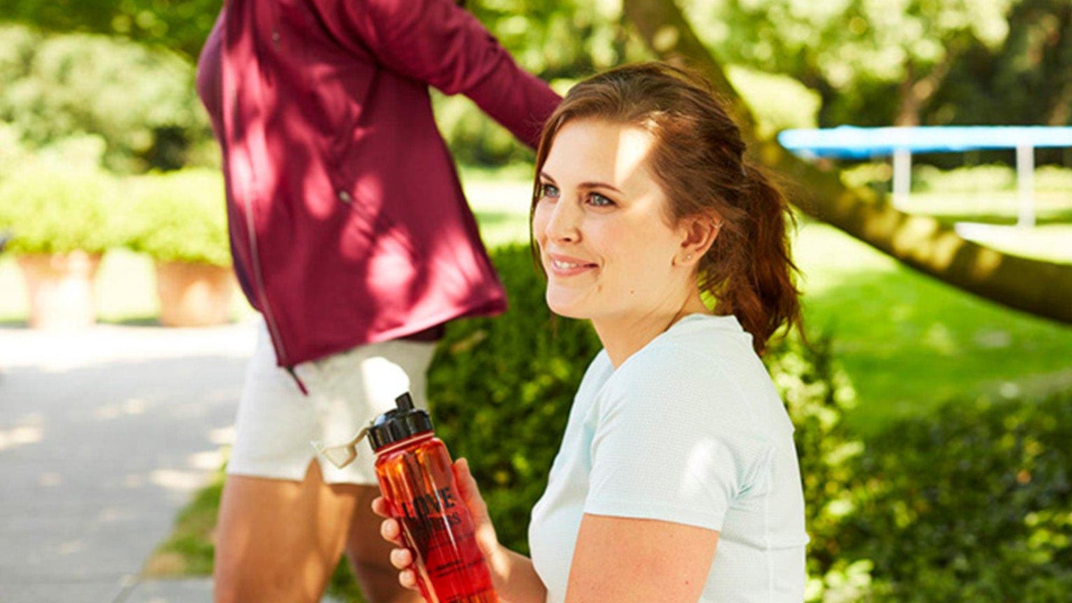 Junge Frau in weissem Sportshirt sitzt mit Trinkflasche auf einer Parkbank.