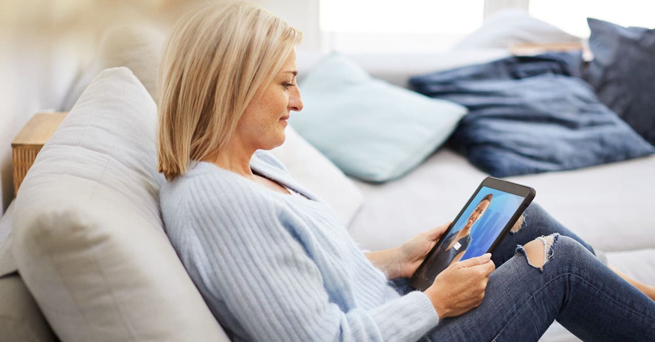 Blonde Frau in hellblauem Pullover sitzt auf Sofa mit Tablet in der Hand.