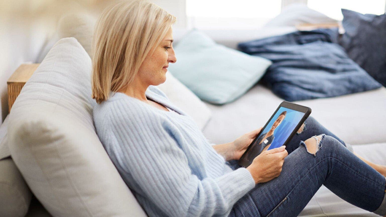 Une femme blonde en pull bleu clair est assise sur un canapé avec une tablette à la main.