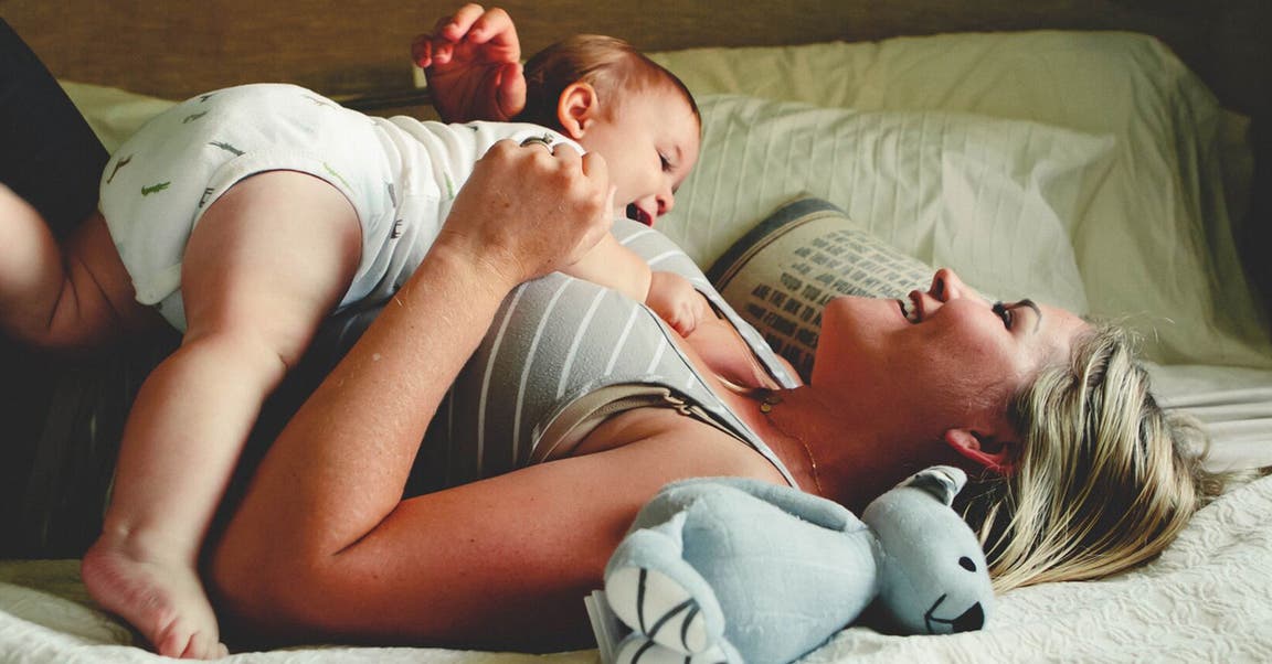 Blondhaarige Frau liegt mit Baby auf einem Bett und lacht.