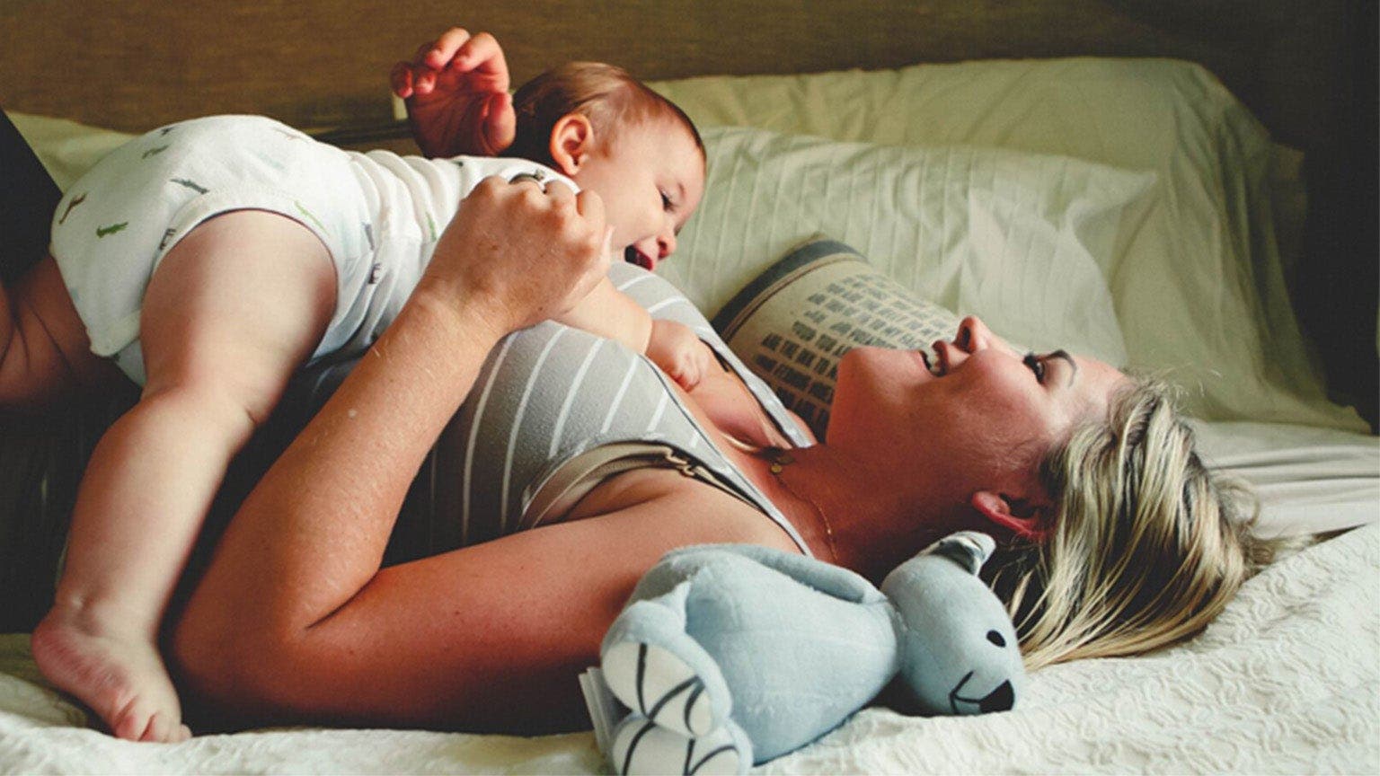 Blondhaarige Frau liegt mit Baby auf einem Bett und lacht.