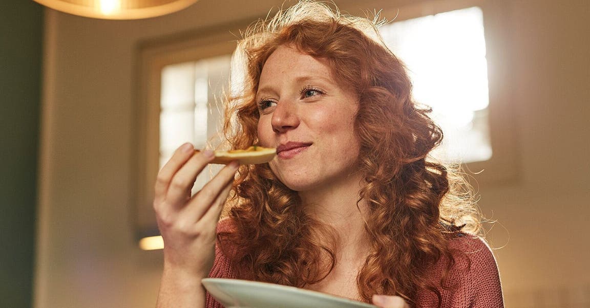 Junge Frau mit roten Haaren isst ein Stück Pizza.