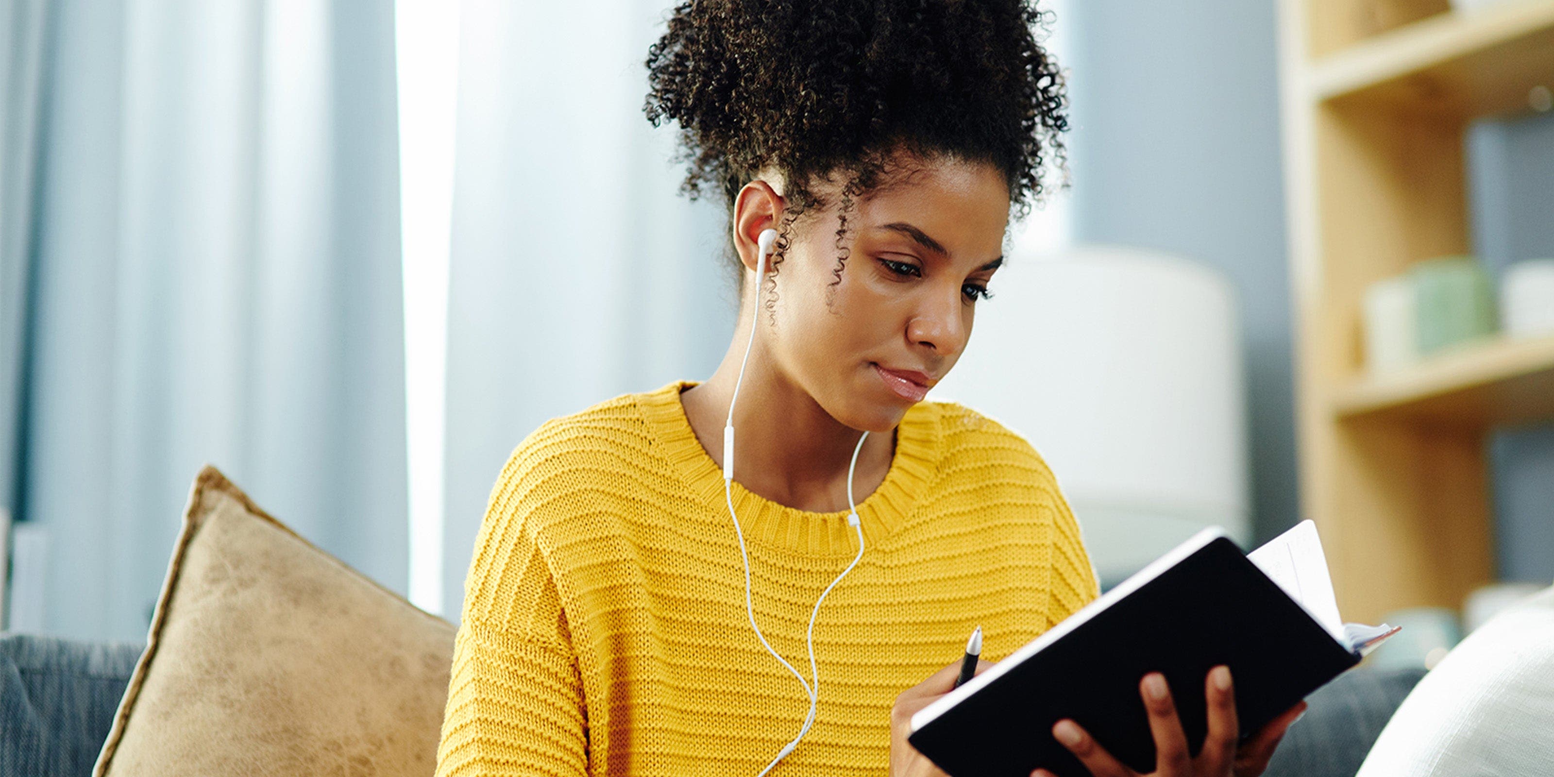 Eine junge Frau sitzt Musik hörend mit einem Notizbuch auf dem Sofa.