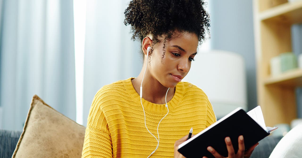 Eine junge Frau sitzt Musik hörend mit einem Notizbuch auf dem Sofa.