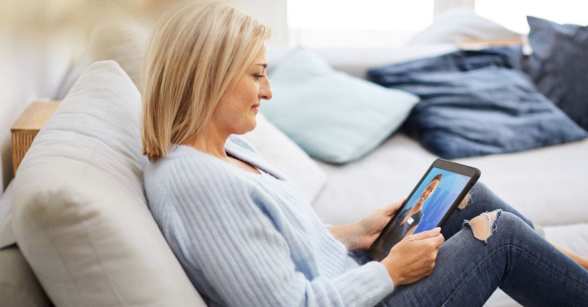 Blonde Frau in hellblauem Pullover sitzt auf Sofa mit Tablet in der Hand.