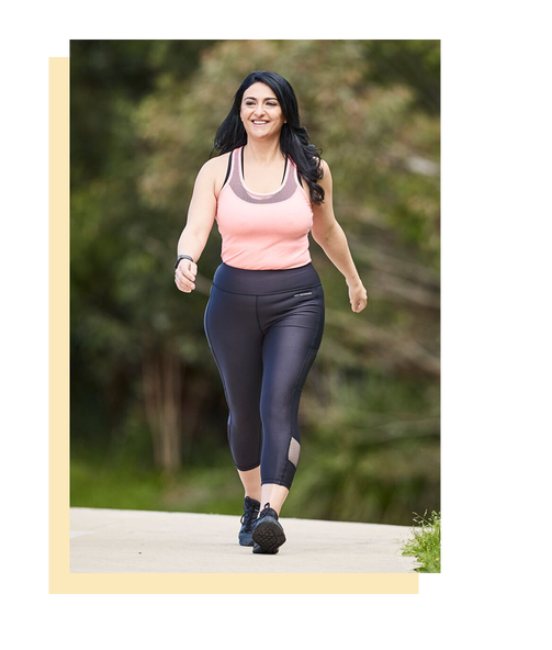 A smiling woman is walking along a path in a park while wearing black pants and sneakers and a pink tank top. Her black hair is cascading over her shoulders.