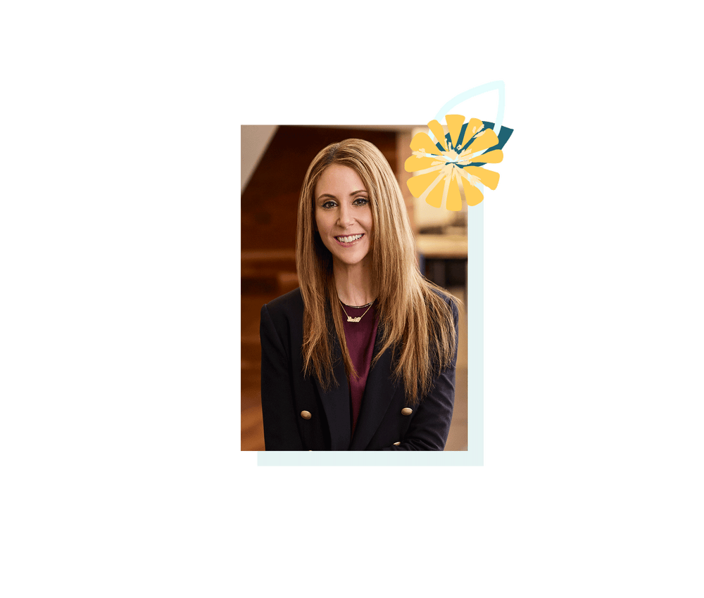 A smiling blonde woman is wearing a dark sport coat with gold buttons and a maroon shirt. Around her neck is a gold necklace.