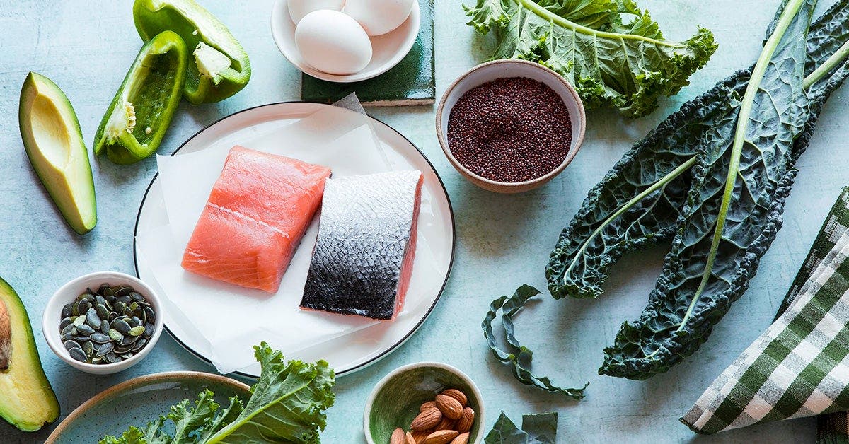 A selection of healthy foods on a kitchen table, including fresh salmon fillets, kale, bell pepper, avocado, eggs, chia seeds, pumpkin seeds, and almonds.