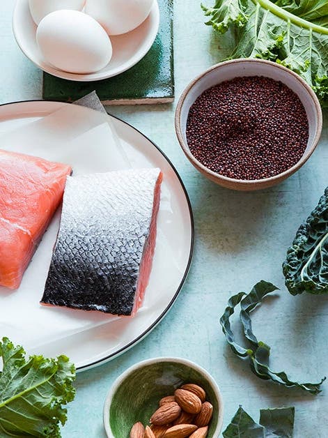 A selection of healthy foods on a kitchen table, including fresh salmon fillets, kale, bell pepper, avocado, eggs, chia seeds, pumpkin seeds, and almonds.