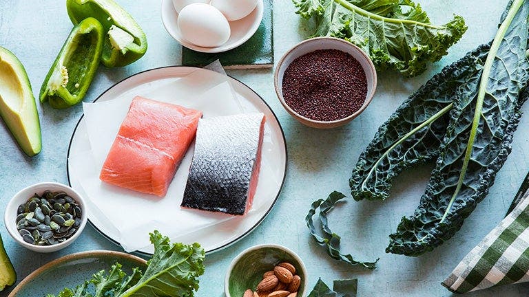 A selection of healthy foods on a kitchen table, including fresh salmon fillets, kale, bell pepper, avocado, eggs, chia seeds, pumpkin seeds, and almonds.