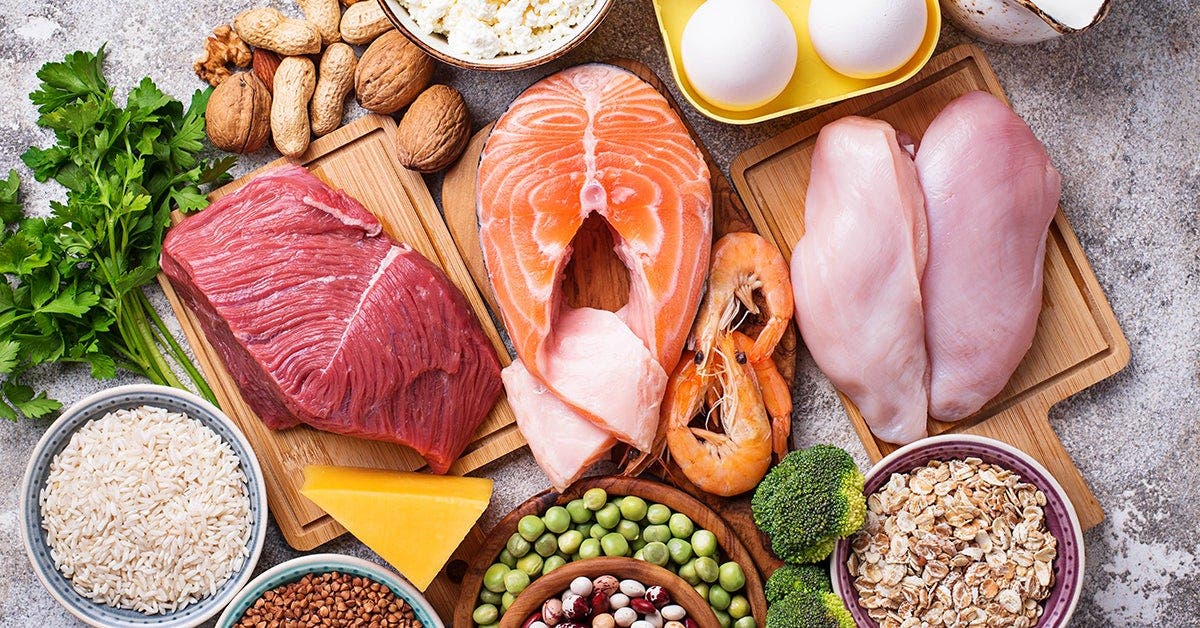 A variety of protein-rich foods including salmon, chicken, beef, eggs, shrimp, cheese, nuts, rice, and beans arranged on a table.