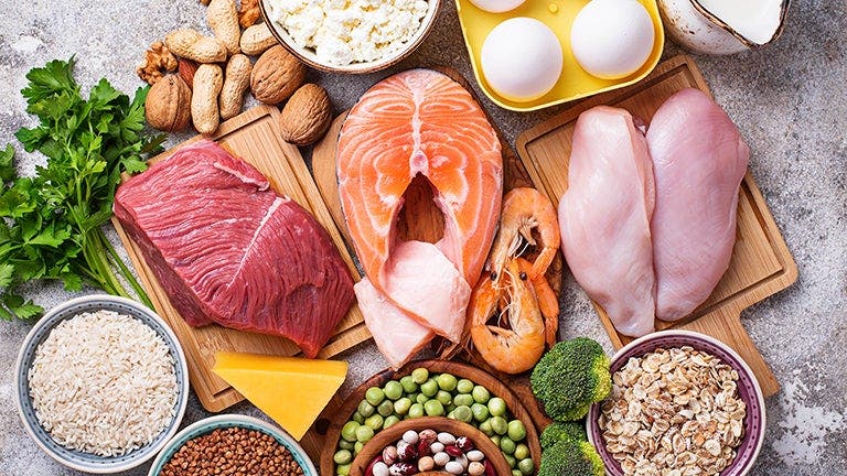 A variety of protein-rich foods including salmon, chicken, beef, eggs, shrimp, cheese, nuts, rice, and beans arranged on a table.