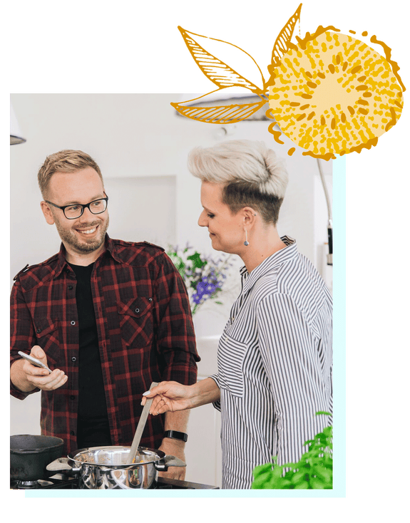 A smiling man with a light beard, blonde hair and black glasses is wearing a black-and-red plaid button-down shirt with a black undershirt and a black wristwatch. He is sharing the screen of his phone with a woman standing to his left. She is wearing a gray-and-white striped shirt and dangly earrings. Her hair is blonde on top, styled in an upward swoosh and is black and very short along the sides and back. They’re both standing in front of a stove and she is stirring a spoon in a silver pot, next to a black pot. There is a green and purple plant in the background.