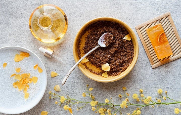 Flat lay of a wooden bowl of coffee-grain body scrub with a spoon, an oil dropper, a bar of soap, and yellow mimosa flowers