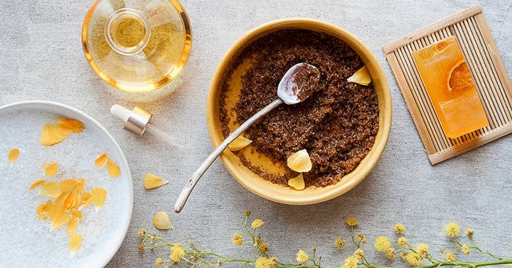 Flat lay of a wooden bowl of coffee-grain body scrub with a spoon, an oil dropper, a bar of soap, and yellow mimosa flowers