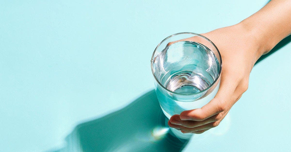 A person&apos;s hand holding a clear glass of water, with a shadow on a turquoise surface.