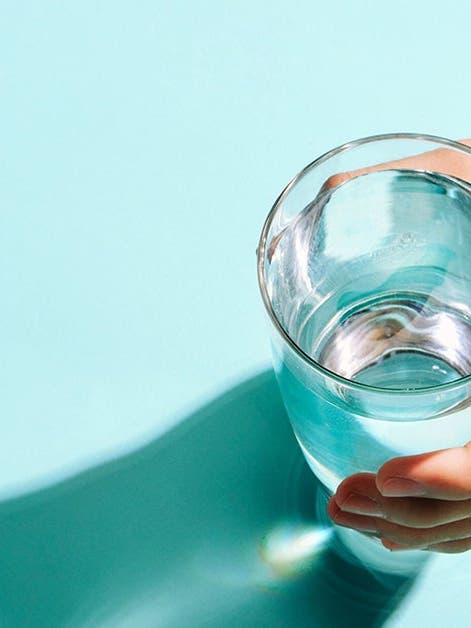A person's hand holding a clear glass of water, with a shadow on a turquoise surface.
