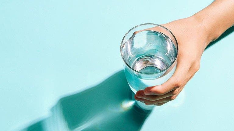 A person&apos;s hand holding a clear glass of water, with a shadow on a turquoise surface.
