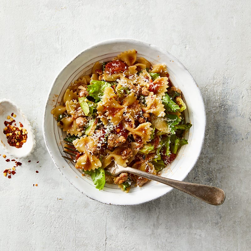 Deep, white serving bowl filled with bow tie pasta with sausage, tomatoes, onions, and escarole, garnished with Parmesan cheese and red pepper flakes
