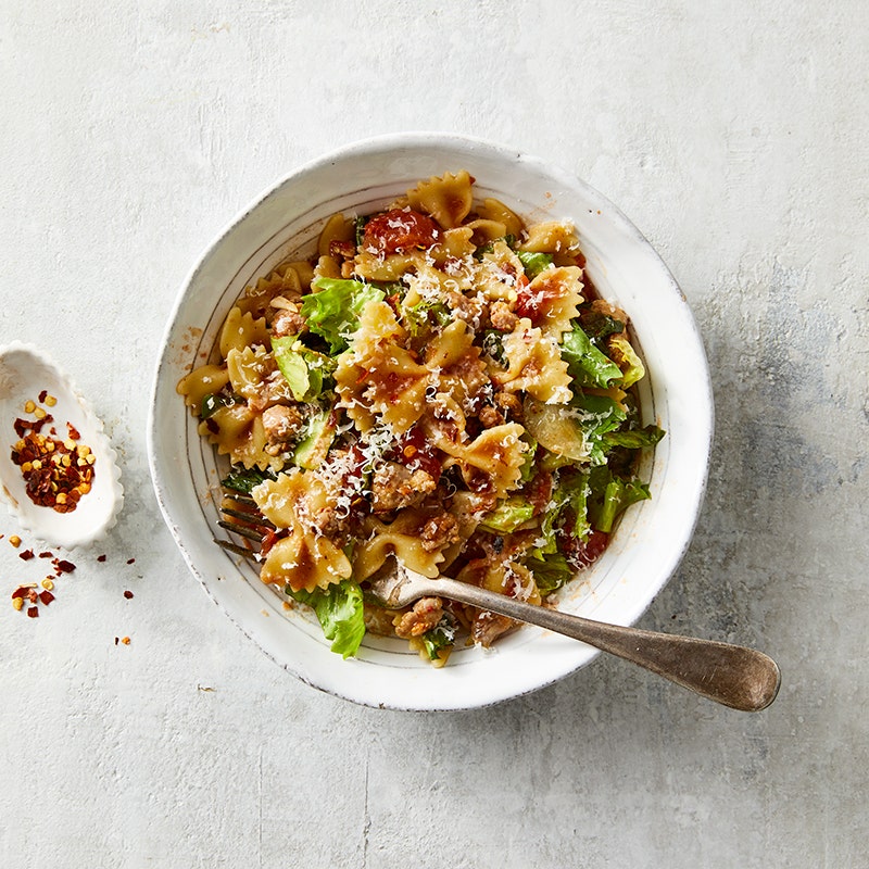 Deep, white serving bowl filled with bow tie pasta with sausage, tomatoes, onions, and escarole, garnished with Parmesan cheese and red pepper flakes
