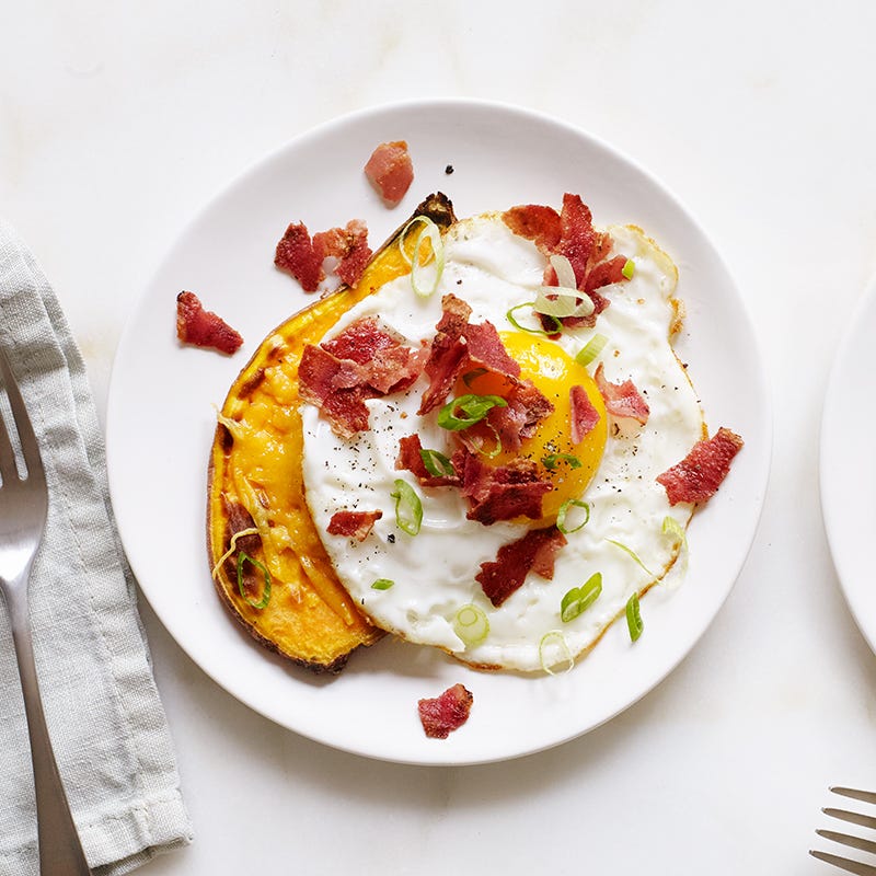 Small round white plate topped with a toasted sweet potato slice, sunny side up egg, crumbled bacon and scallions