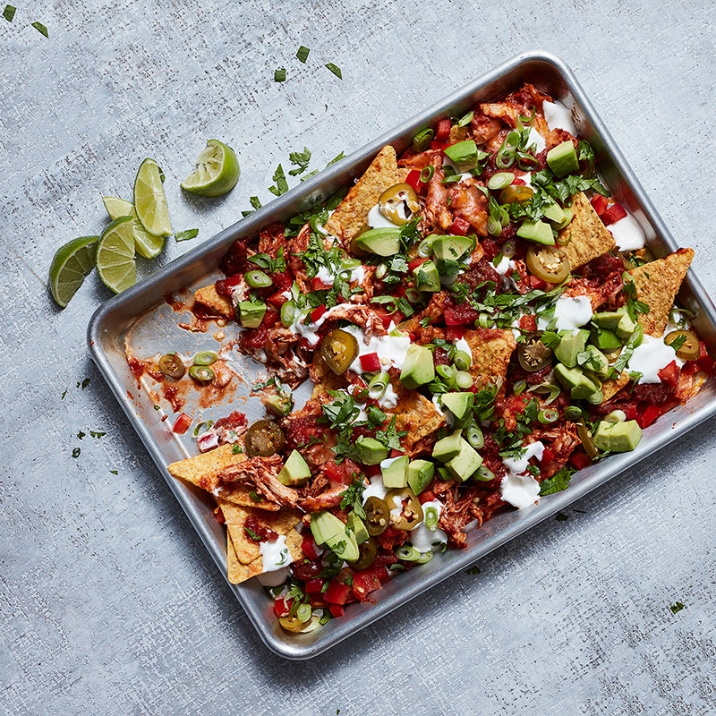 a sheet pan filled with pulled chicken-topped tortilla chips that are garnished with cheese, salsa, avocado, and fresh vegetables
