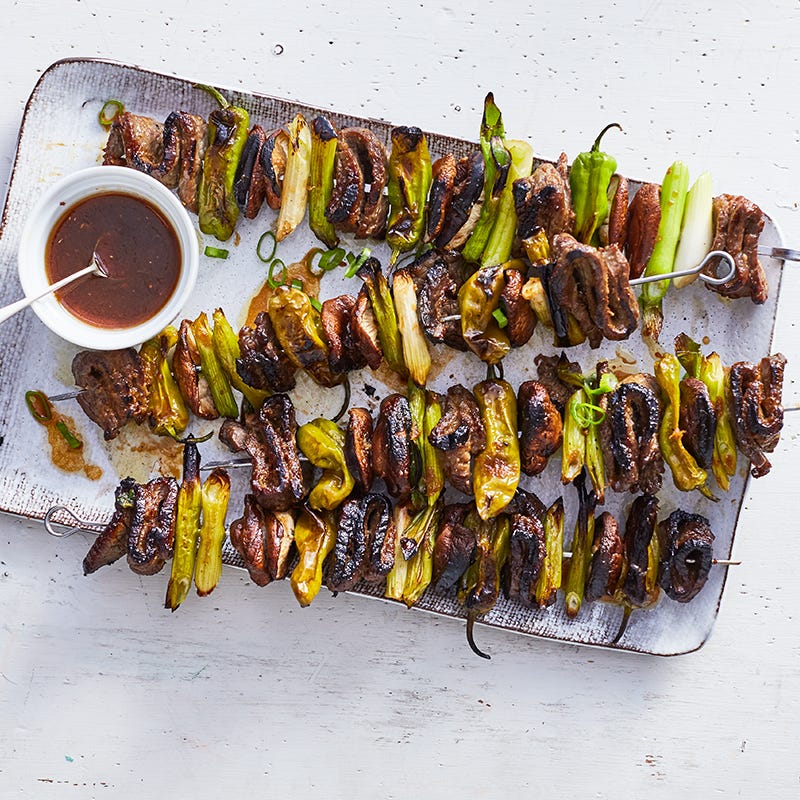 Rectangular serving platter topped with skewers of grilled beef, mushrooms, and peppers, with a small bowl of dipping sauce