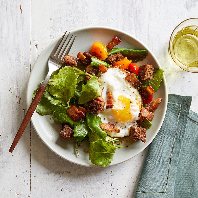 Round white plate topped with kale, tomatoes, chopped bacon, croutons and a sunny-side up egg