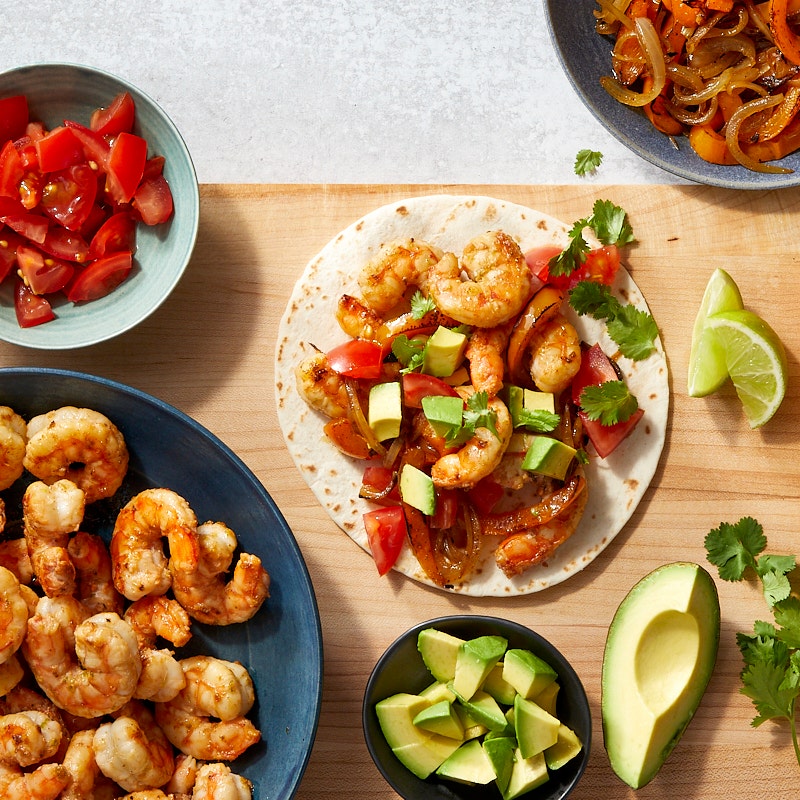 a wooden surface with individual bowls of cooked shrimp, cooked peppers and onions, chopped tomatoes, and chopped avocado with an already prepped fajita on the surface