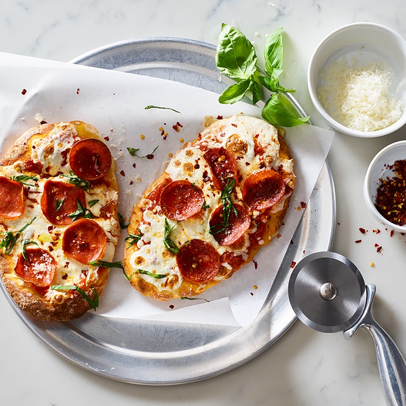 Two individual turkey pepperoni pizzas on a circular pizza pan with Parmesan cheese, basil and pepper flakes on the side