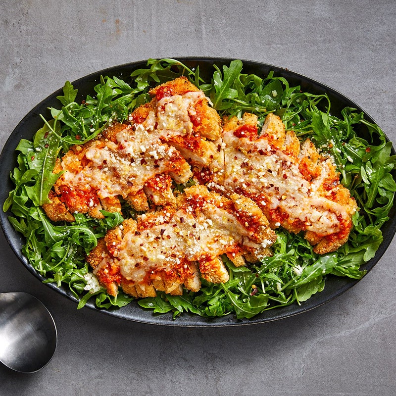 large, oval black bowl filled with arugula, tossed with a vinaigrette, and topped with sliced, air-fried chicken Paremsan