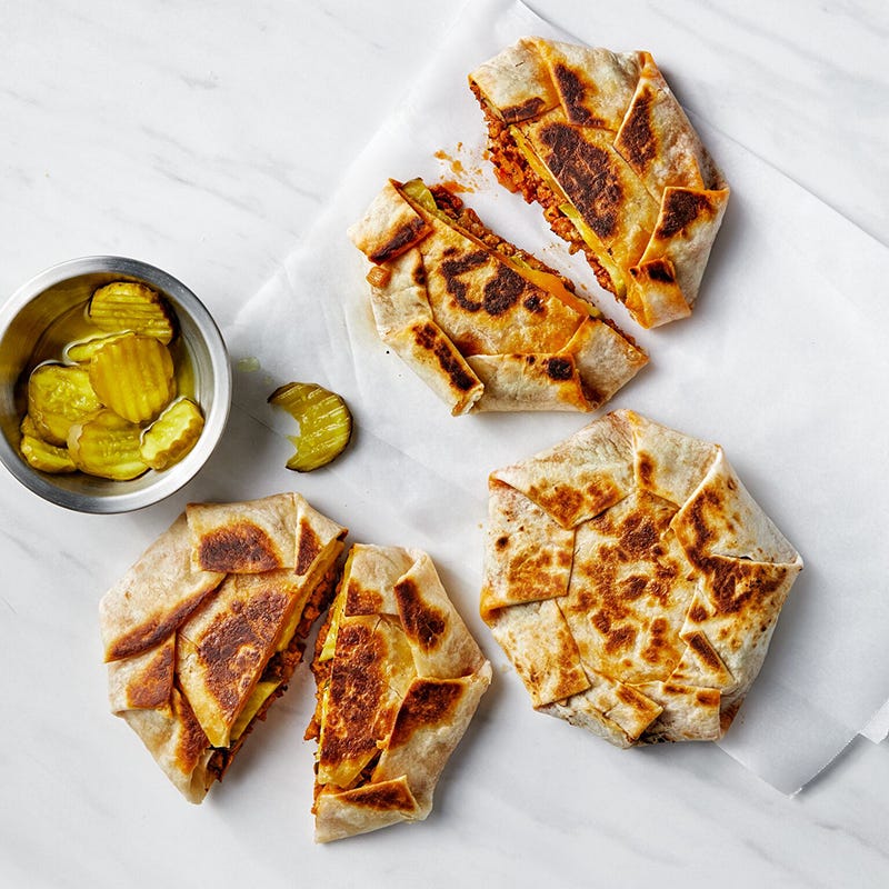 image of 3 sloppy Joe crunch wraps on parchment paper with a small bowl of pickles on the side