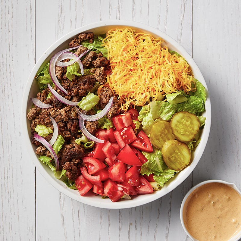 White bowl filled with lettuce and topped with sauteed ground beef, chopped tomatoes, shredded cheese, and pickles, with a small bowl of dressing on the side
