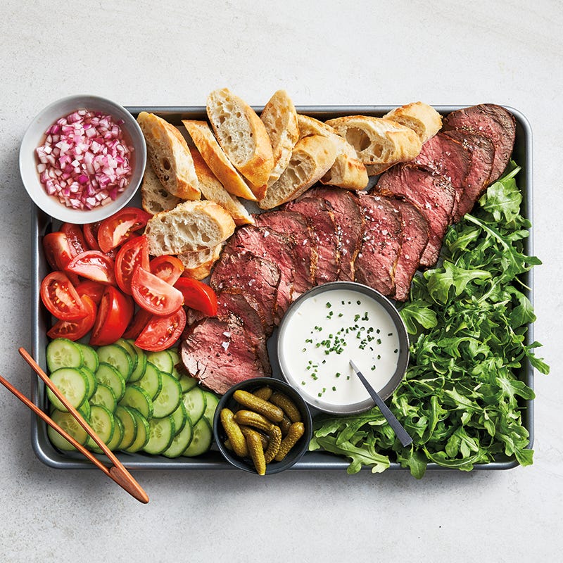 Large sheet pan topped with sliced baguette, beef, cucumber and tomato, baby arugula, and small bowls with minced onion, cornichon, and a creamy horseradish sauce