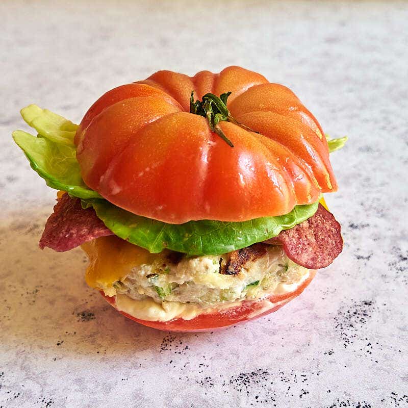 A turkey cheeseburger topped with turkey bacon, lettuce, and mayonnaise, sandwiched between 2 tomato halves as a bun