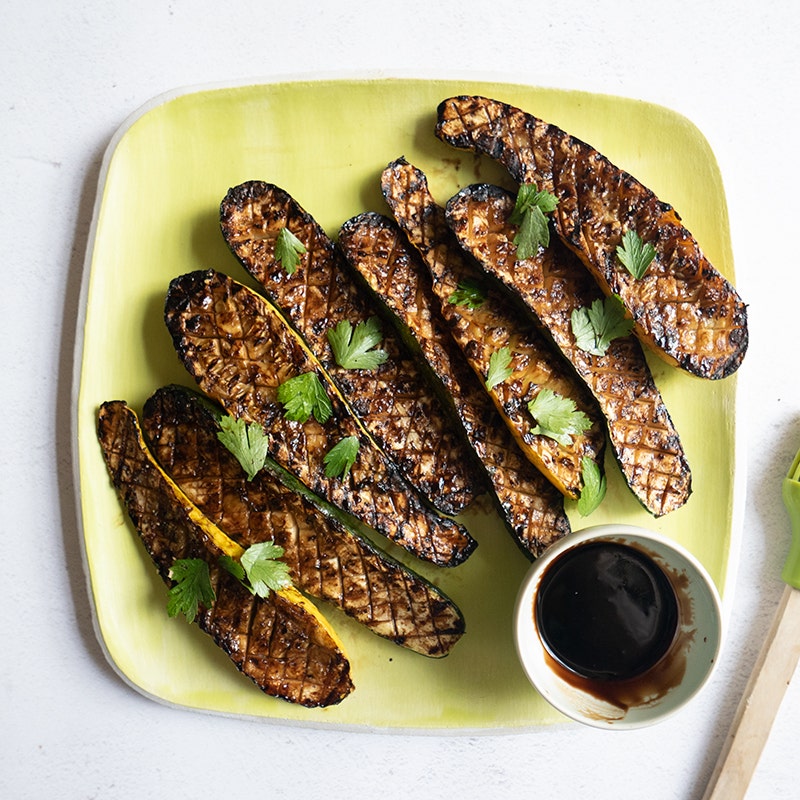Square serving platter topped with grilled summer squash halves, served with a small dish of balsamic glaze
