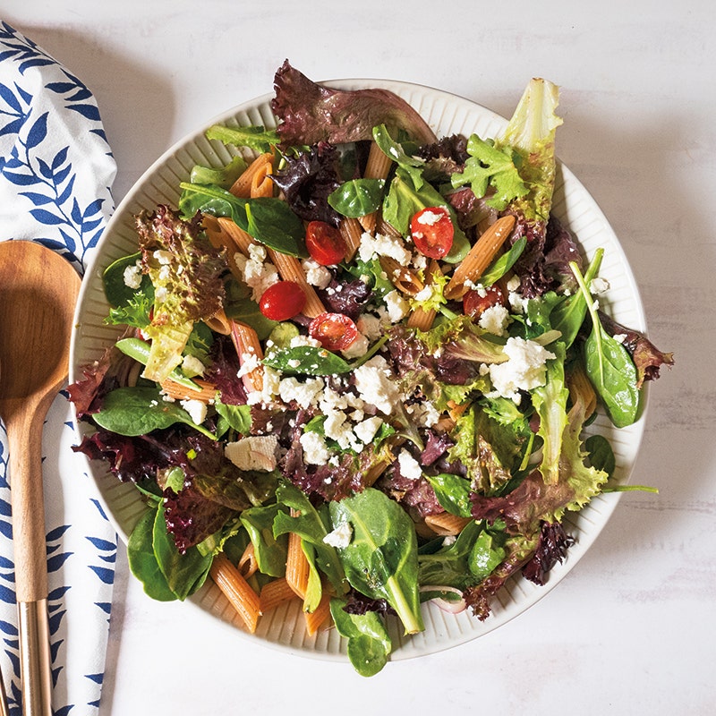large round serving dish filled with a mixture of penne pasta salad made with tomatoes, onions, feta, and mixed greens
