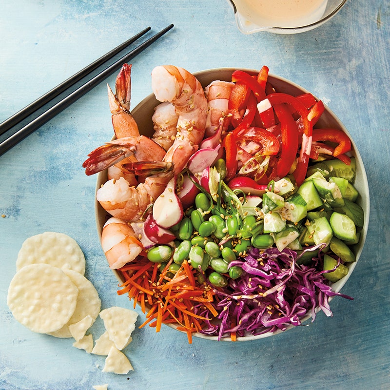 A large bowl with sections of cabbage, carrot, bell peppers, edamame, cucumber, radishes and shrimp served with a spicy sesame drizzle and garnished with sesame seeds, scallions and rice crackers.